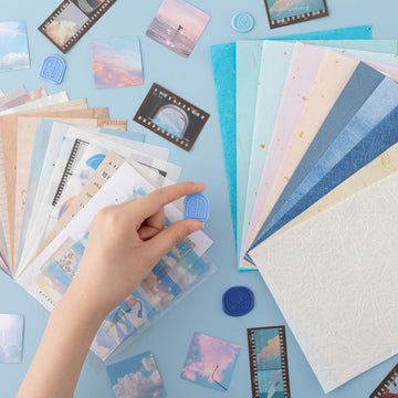 Hand holding a wax seal stamp from the Tsuki ‘Cloud Dreamland’ Scrapbooking Set with paper and stickers spread out in the background